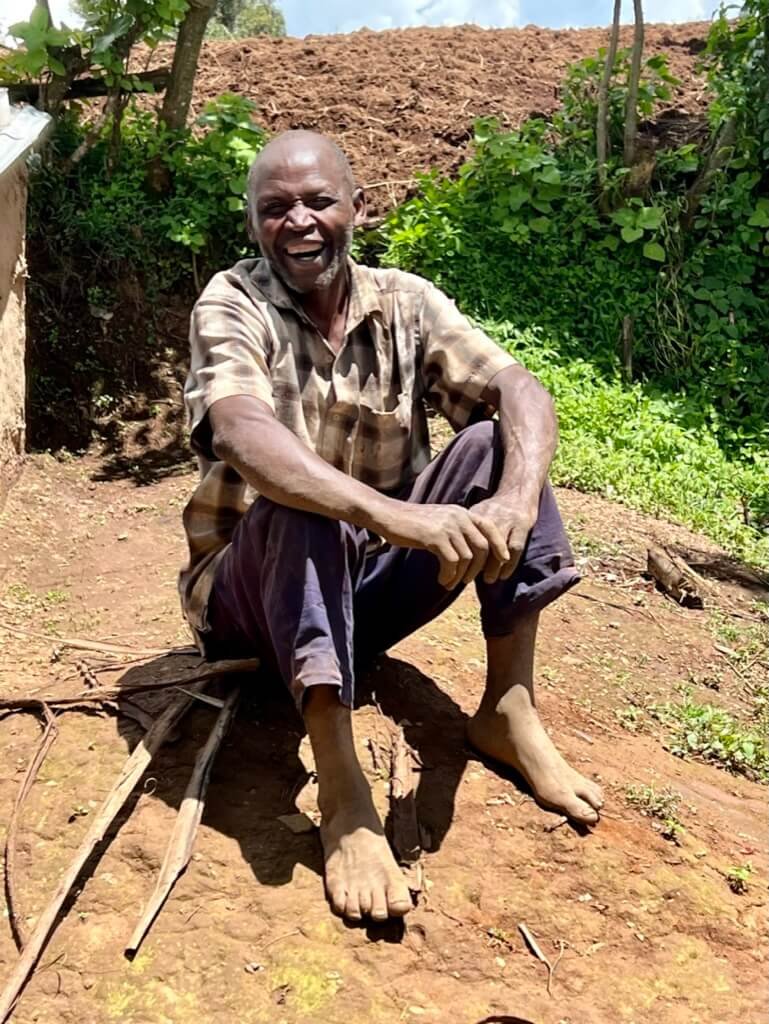 An elderly man sits barefoot on the ground, smiling widely. He is wearing a plaid shirt and dark pants. The background features greenery and a sloped, earthy terrain under a clear sky. Uganda Amatsiko Tours