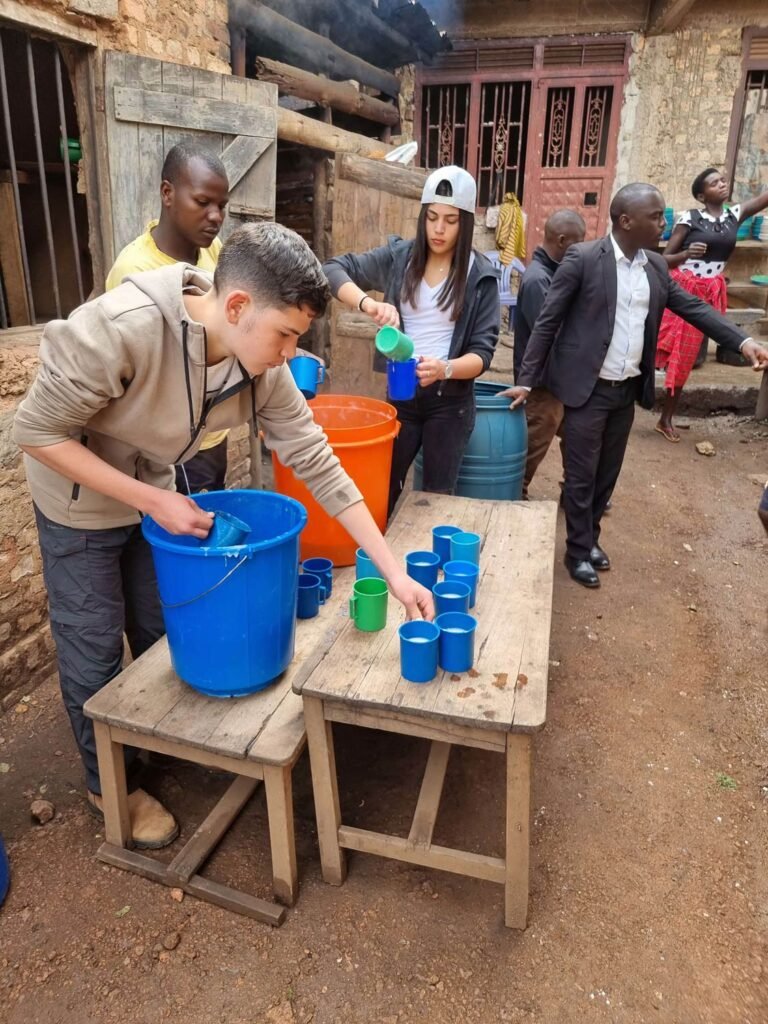 A group of people pour water from large orange and blue containers into smaller colorful cups on wooden tables outdoors. They are in a rustic setting with brick walls and wooden doors. Uganda Amatsiko Tours