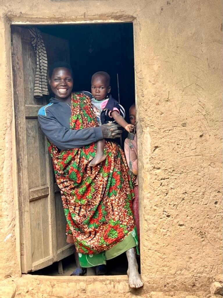 A person in colorful clothing stands in a doorway holding a child. The doorway is part of a rustic building with earth-toned walls. Another child peeks from behind them. The scene conveys a sense of warmth and family. Uganda Amatsiko Tours