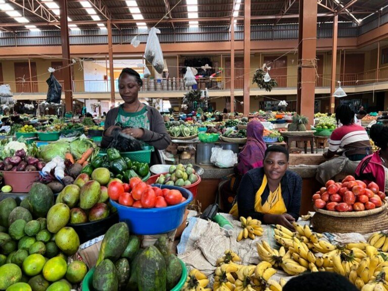 A bustling indoor market with vendors selling fresh produce. In the foreground, there are colorful piles of mangoes, bananas, tomatoes, and avocados. Two vendors are smiling and interacting with customers. The market is lively and vibrant. Uganda Amatsiko Tours