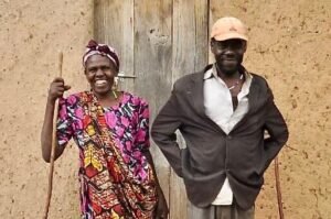 A smiling woman and man stand outside a rustic building with a wooden door. The woman wears a colorful dress and headscarf, holding a stick. The man wears a cap, suit jacket, and holds his hands behind his back. Uganda Amatsiko Tours