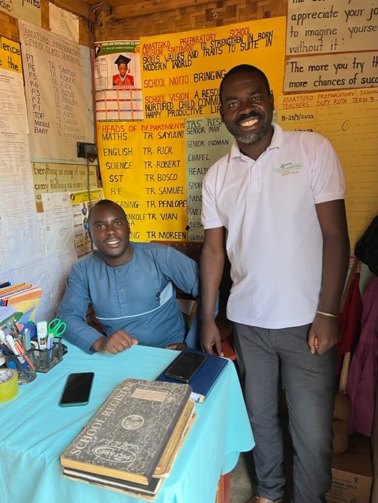 Two men smiling in an office with educational posters on the walls. One is seated at a desk with books and stationery, while the other stands beside him wearing a white polo shirt. Uganda Amatsiko Tours