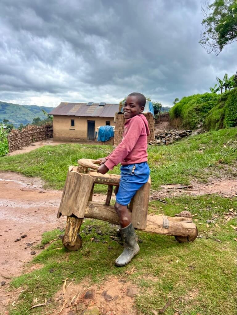 A smiling child stands outside on a homemade wooden scooter on a dirt path. They wear a pink sweater, blue shorts, and rain boots. In the background are a house, greenery, and a cloudy sky. Uganda Amatsiko Tours