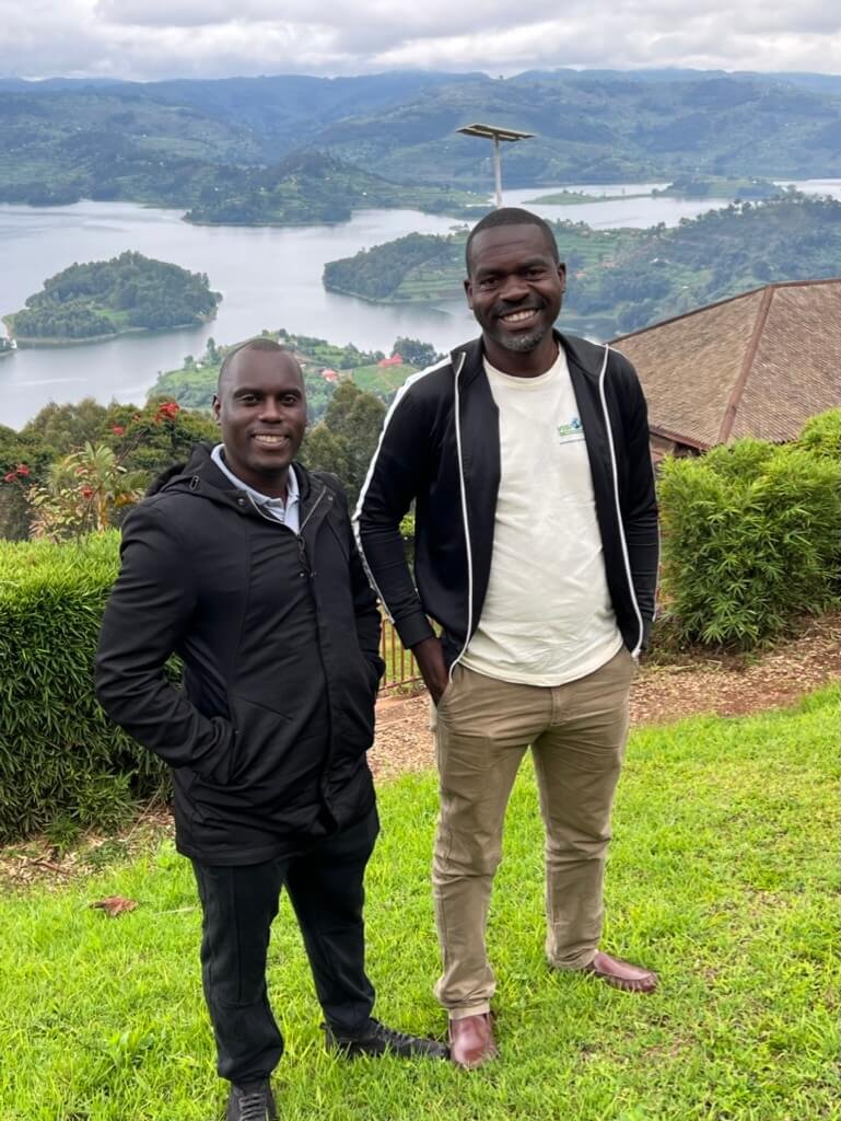 Two people are standing on a grassy area, smiling at the camera. Behind them is a picturesque view of a lake surrounded by hills and greenery on a cloudy day. They are wearing casual clothing, with jackets and trousers. Uganda Amatsiko Tours