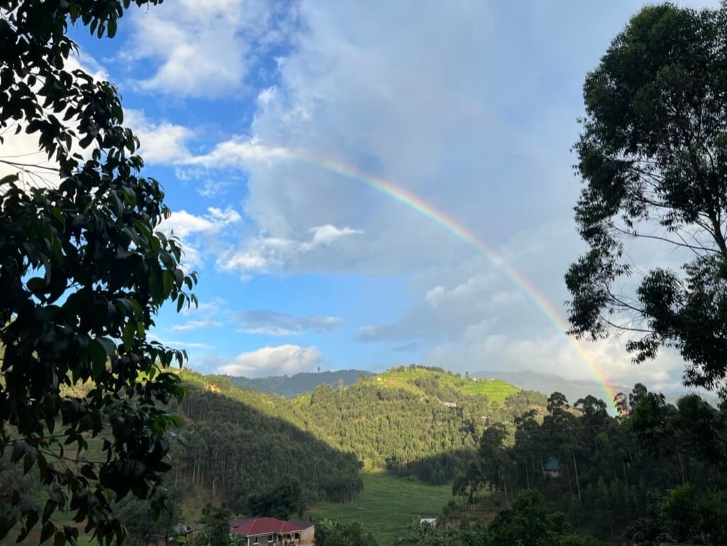 A lush green valley surrounded by forested hills under a partly cloudy blue sky. A vibrant rainbow arches across the scene, with a small house partially visible in the foreground. Uganda Amatsiko Tours