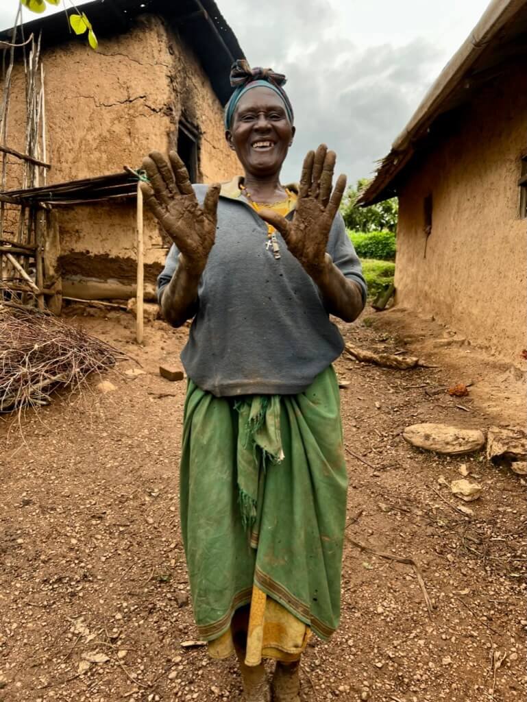 A smiling woman stands outdoors with her hands covered in mud, showing them to the camera. She is wearing a headscarf, a gray top, and a green skirt, and is surrounded by rustic, mud-brick structures. The sky is cloudy. Uganda Amatsiko Tours