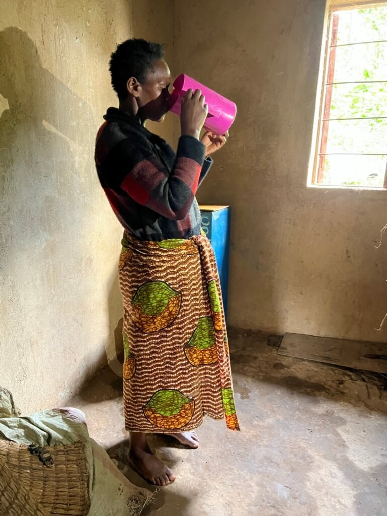 A person stands indoors, drinking from a pink cup. They wear a black and red sweater and a patterned skirt. The room has a window with bars, and the floor is concrete. Sunlight filters in, creating a warm atmosphere. Uganda Amatsiko Tours