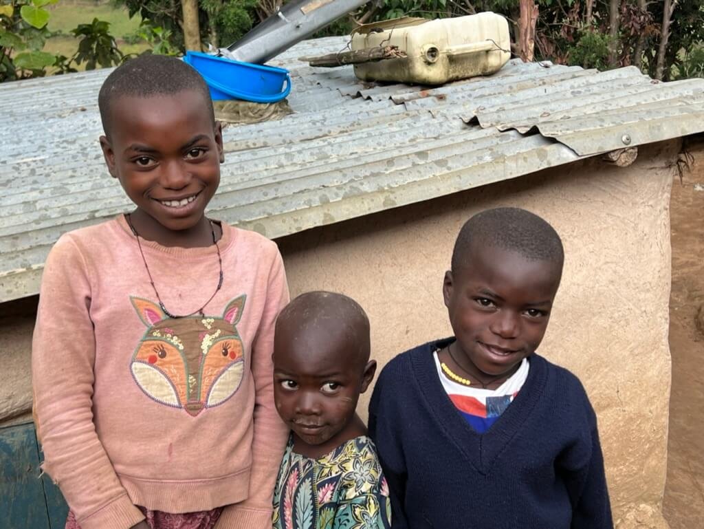 Three children stand smiling in front of a small, corrugated metal-roofed building. One wears a pink sweater with a fox design, another is in floral clothing, and the third wears a dark sweater. A plastic container and basin are on the roof. Uganda Amatsiko Tours