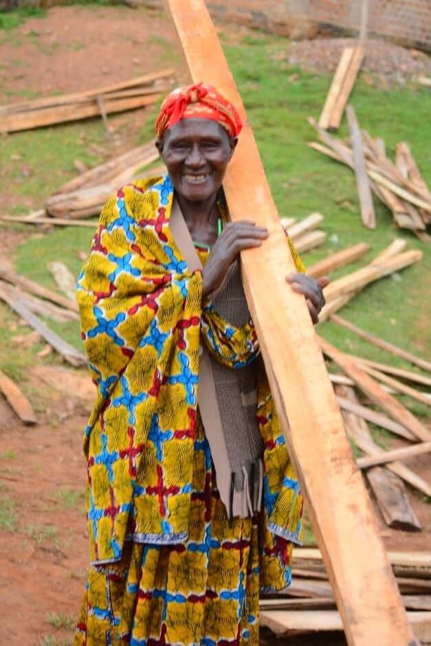 An elderly person wearing colorful traditional clothing and a red headscarf smiles while carrying a wooden plank. There are more wood planks scattered on the ground in the background. Uganda Amatsiko Tours