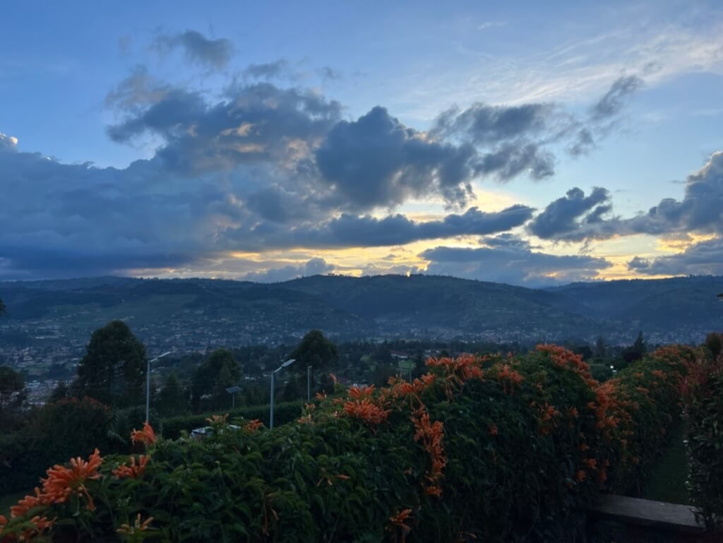 A scenic view of a hilly landscape at sunset, with a sky filled with dramatic clouds. Orange flowers line the foreground, adding vibrant color. The distant hills and scattered urban lights suggest a peaceful, rural atmosphere. Uganda Amatsiko Tours