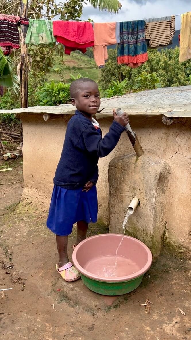 A young child in a blue skirt and dark sweater stands by a water spout, filling a basin. The background shows colorful clothes drying on a line and lush greenery. Uganda Amatsiko Tours