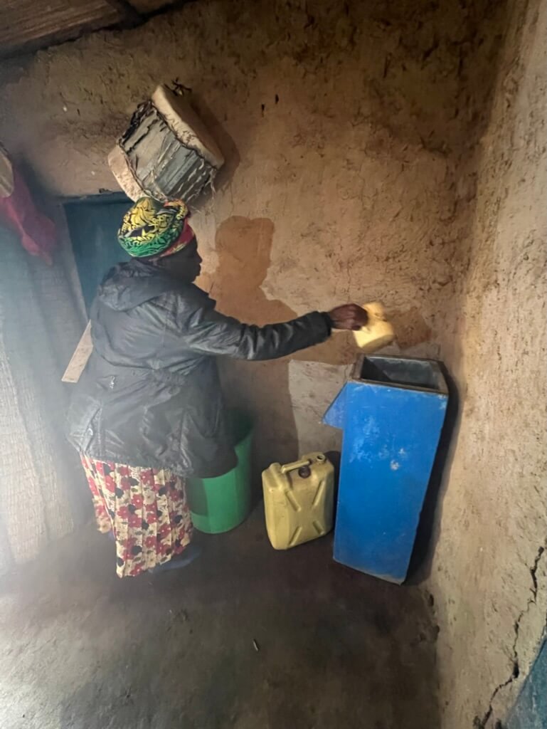 A person wearing a colorful headscarf and patterned skirt is in a dimly lit room, placing a plastic container into a blue box. Yellow jerry cans are on the ground nearby, and traditional baskets hang on the wall. Uganda Amatsiko Tours