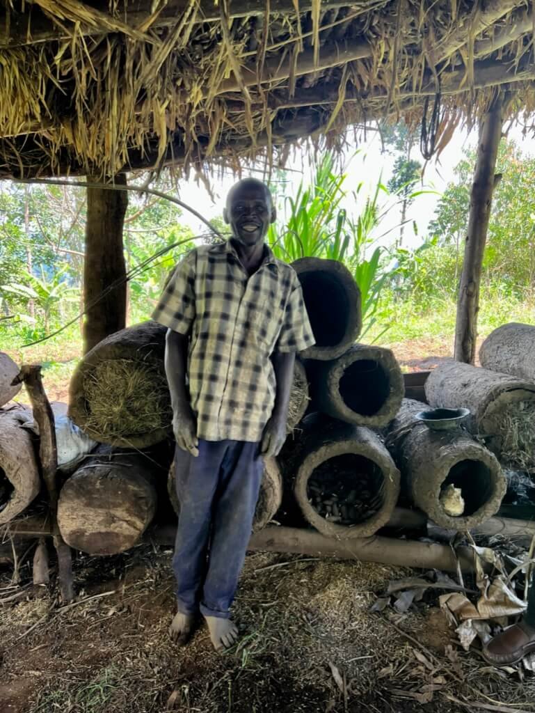 A person stands smiling under a thatched roof next to large hollow logs stacked on the ground. Lush greenery is visible in the background. The person is barefoot and wearing a checkered shirt and dark pants. Uganda Amatsiko Tours