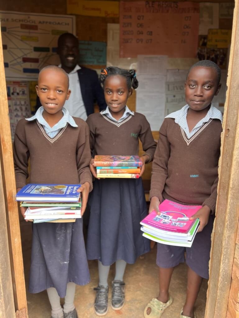 Three children in school uniforms stand in a classroom doorway, each holding a stack of books. They are smiling, and a man in the background is also smiling. Educational posters are visible on the walls. Uganda Amatsiko Tours