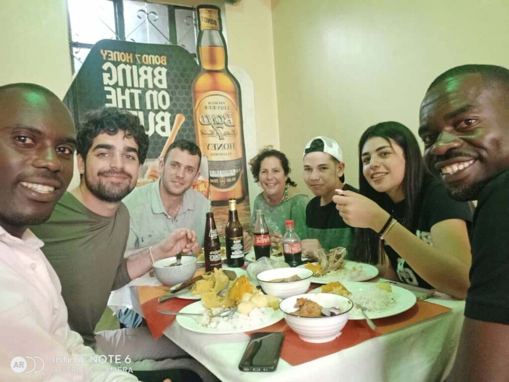 A group of six people sitting at a dining table, enjoying a meal together. There are various dishes and drinks on the table. A poster of a whiskey bottle is visible in the background. Everyone is smiling at the camera. Uganda Amatsiko Tours