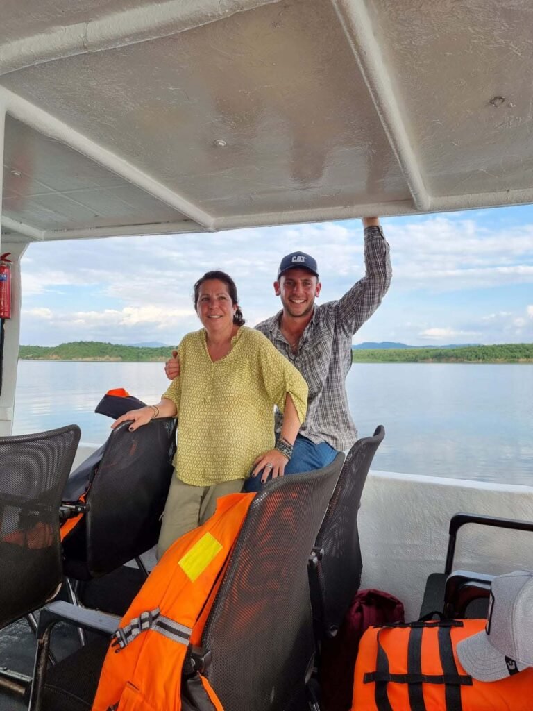 Two people smiling on a boat, with a lake and trees visible in the background. They are surrounded by life jackets and outdoor gear, suggesting an adventure or excursion. The man is wearing a cap and leaning casually against the boat's roof. Uganda Amatsiko Tours