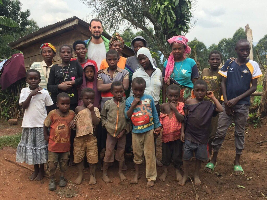 A group of people, including children and adults, stand together outside on a dirt ground with trees and a small building in the background. They are dressed in casual clothing, some smiling and some with neutral expressions. Uganda Amatsiko Tours