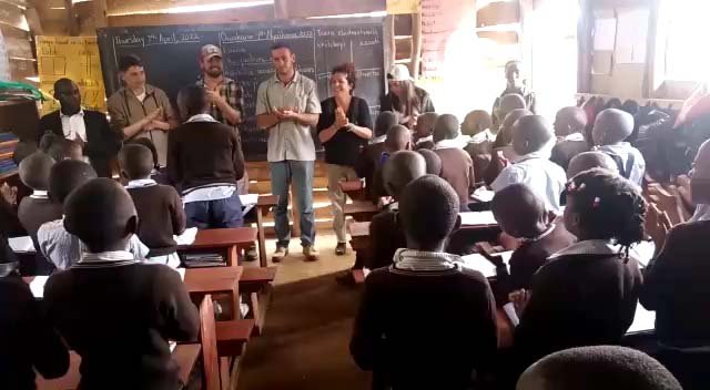 A group of adults stands at the front of a classroom, clapping. They are in front of a blackboard. Children in brown uniforms are seated at wooden desks, facing them and participating in the activity. Uganda Amatsiko Tours