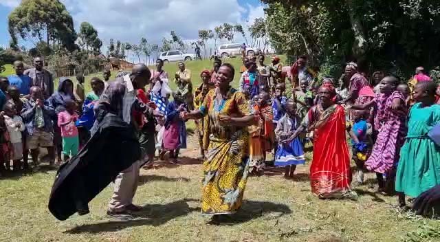 A lively outdoor celebration with people dancing in colorful traditional attire. Children and adults gather around on a grassy field under a partly cloudy sky, with parked cars in the background. Uganda Amatsiko Tours