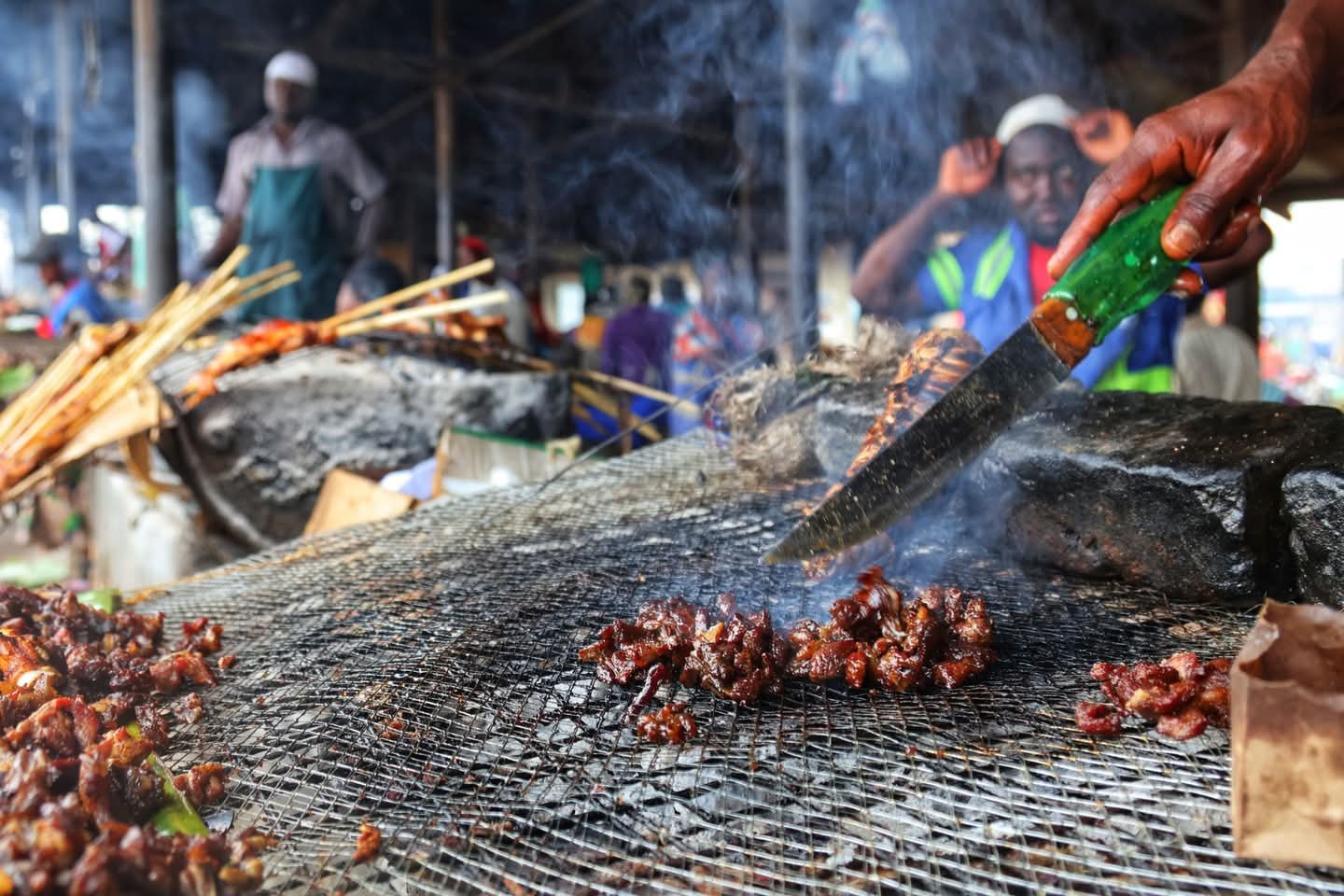 Meat being roasted on a wire mesh and someone holding a knife to cut the meat.